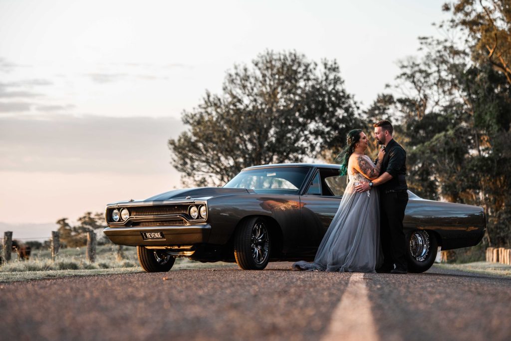 A couple in formal attire standing close and embracing next to a vintage black car on a rural road at sunset with trees in the background.