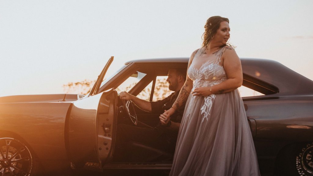 A woman in a gray dress holding hands with a man in the driver's seat of a black vintage car during sunset.