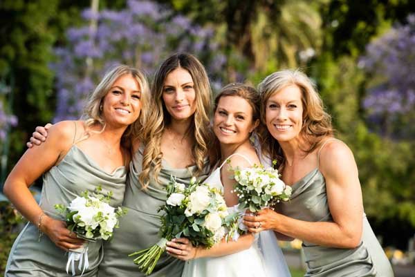 Four women in gray dresses holding bouquets of white flowers, smiling outdoors with trees and purple blooming flowers in the background.