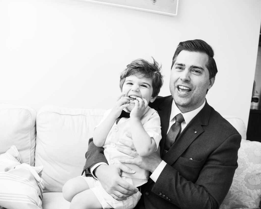 A man in a suit holding a laughing young boy on a sofa in a living room.
