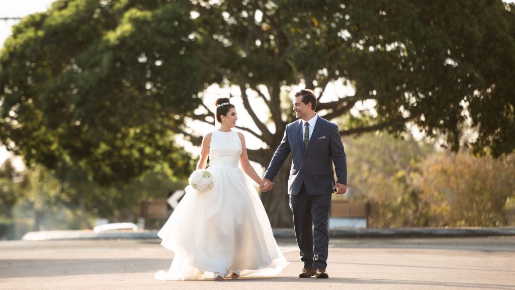 A bride and groom walking hand in hand outdoors, smiling at each other on their wedding day, with large trees in the background.