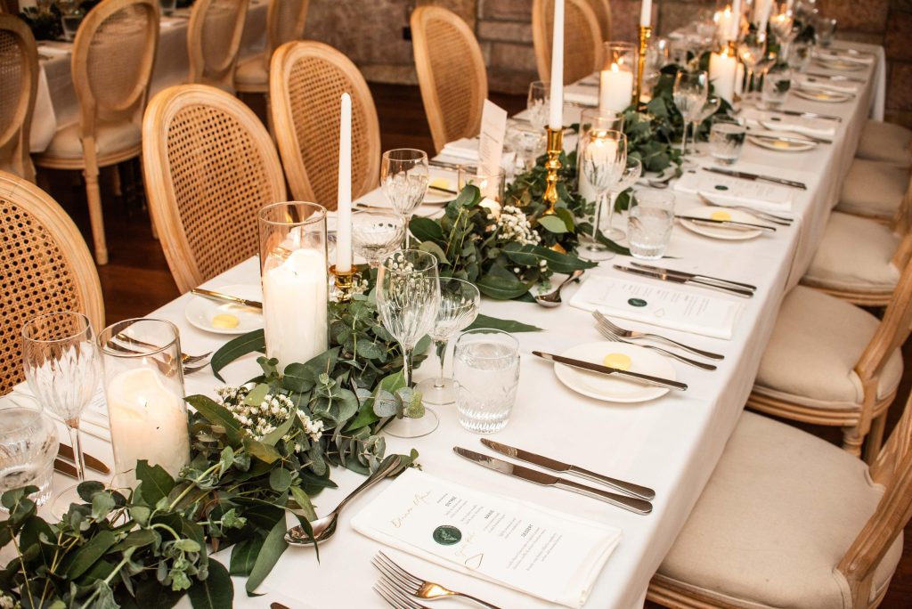 Elegant dining table set with candles, greenery, gold candle holders, wine glasses, and place settings with napkins and cutlery.