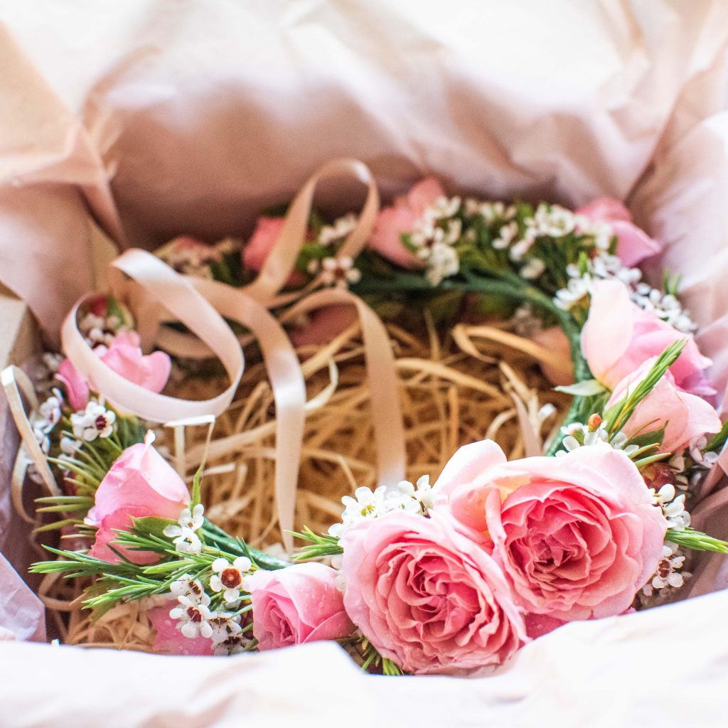 Pink and white floral wreath with roses, small white flowers, and green leaves placed inside a cardboard box with tissue paper