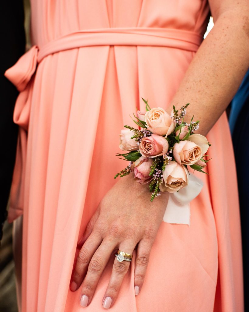 Close-up of a woman's hand with wedding rings and a floral corsage on her wrist, wearing a peach-colored dress with pleats.