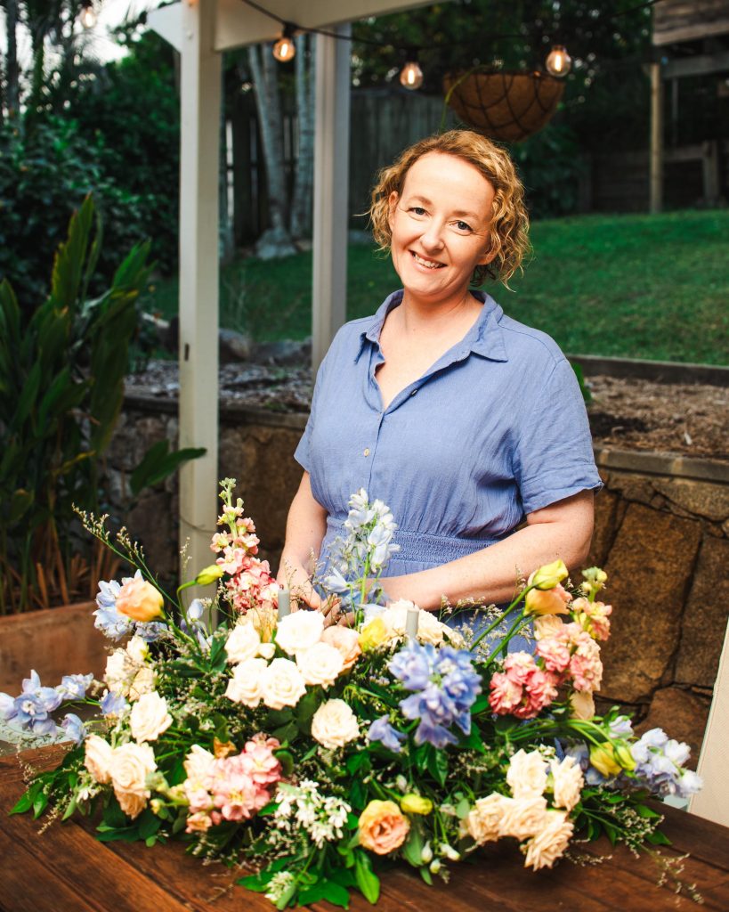 A woman with curly red hair wearing a blue dress stands behind a large bouquet of colorful flowers on a wooden table outdoors, with a garden and string lights in the background.