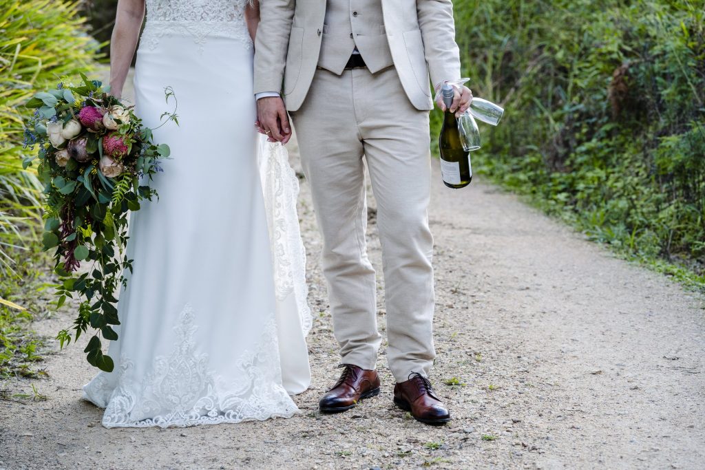 A bride and groom holding hands, with the bride holding a bouquet, standing on a dirt path surrounded by greenery.