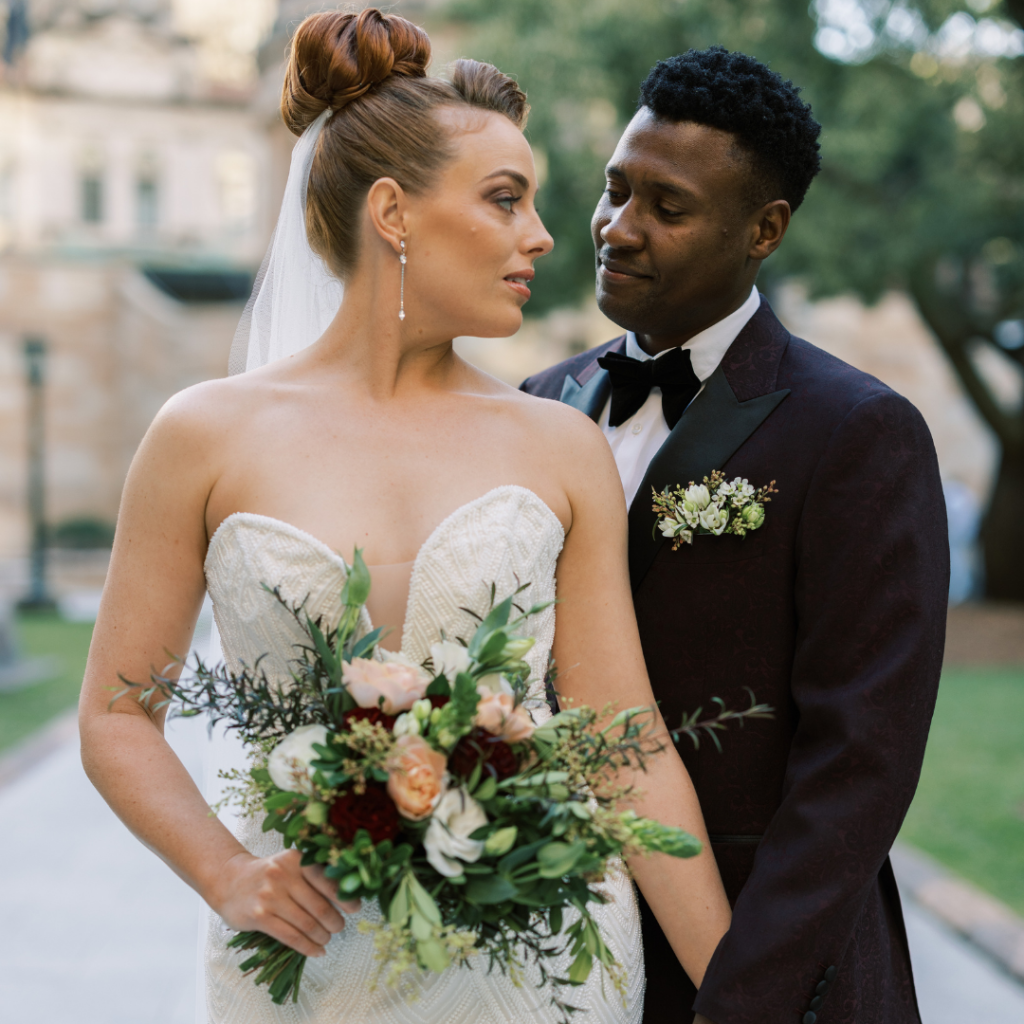 A bride in a white strapless wedding dress holding a bouquet looking into the eyes of a groom in a dark tuxedo with a boutonniere, outdoors on a wedding day.