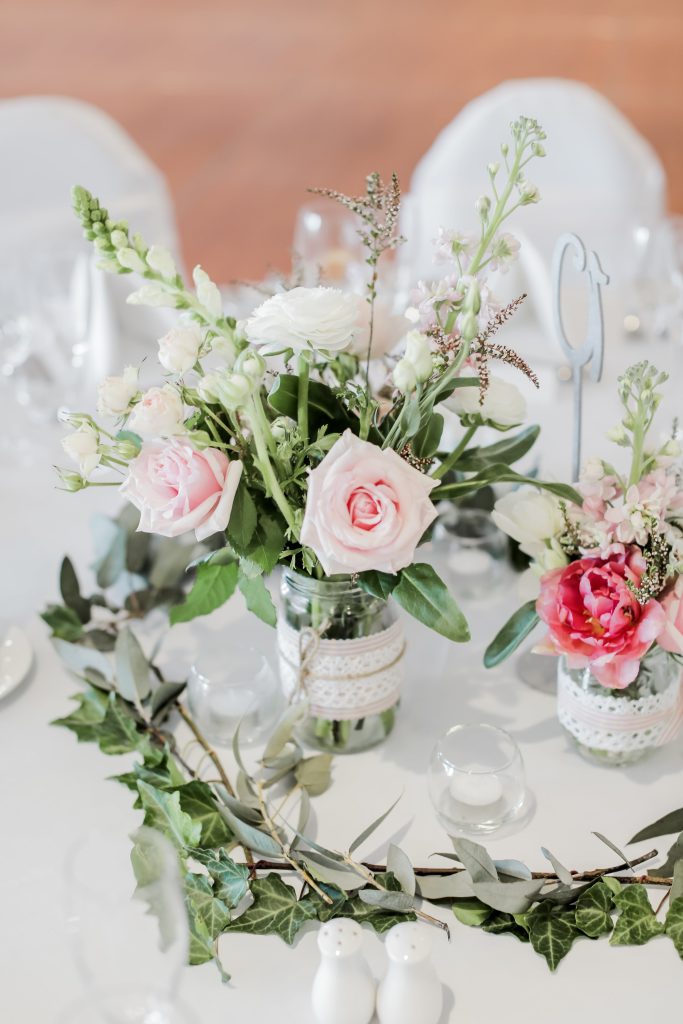 A charming and rustic wedding table setting featuring mason jar centerpieces filled with a vibrant mix of pastel flowers. The arrangement includes roses, lisianthus, and other seasonal blooms, creating a cheerful and inviting atmosphere for the guest