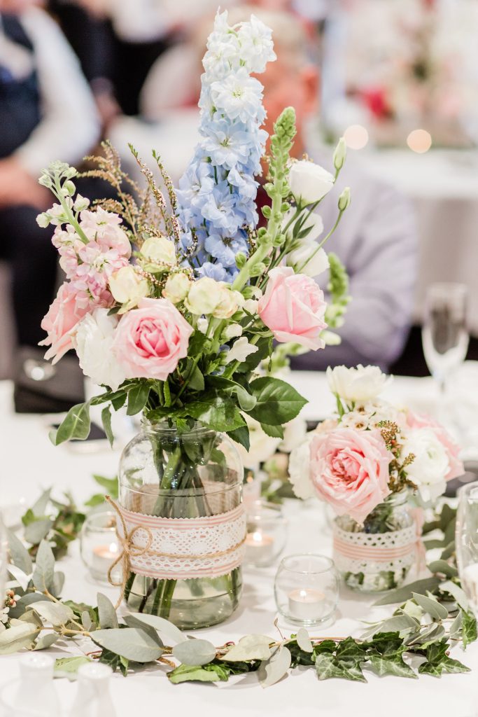 A charming and rustic wedding table setting design by Hart & Flowers featuring mason jar centerpieces filled with a vibrant mix of pastel flowers. The arrangement includes roses, lisianthus, delphinium and other seasonal blooms, creating a cheerful a