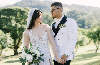 A smiling bride in a lace gown and veil holds hands with her groom in a white tuxedo at Austinvilla Estate. The bride is holding an elegant, natural-style bouquet featuring white dahlia and eucalyptus. The groom has a modern pocket buttonhole in his