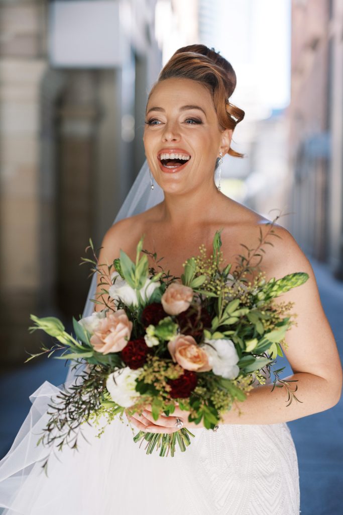 A smiling bride with red hair holding a bouquet of pink, white, and burgundy flowers, wearing a white wedding dress and earrings.