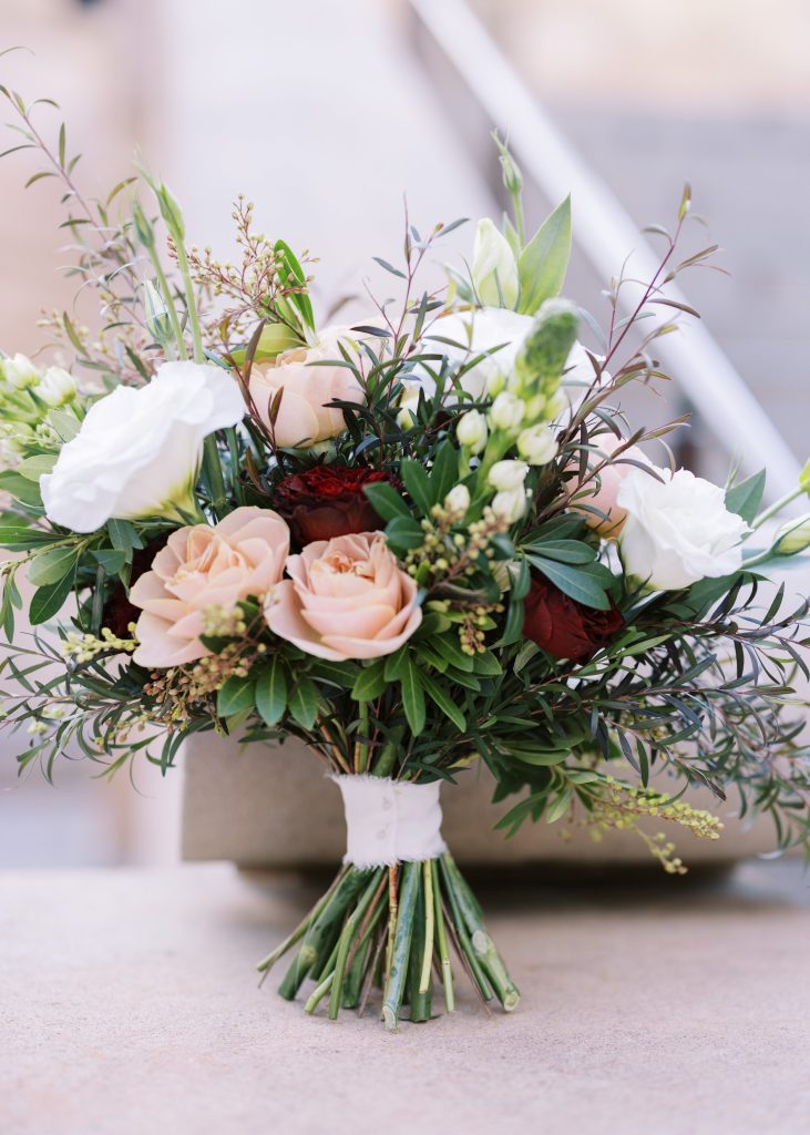 A bouquet of mixed flowers including pale pink roses, white lisianthus, and dark red blooms with green foliage, tied with white cloth, resting on a beige surface.