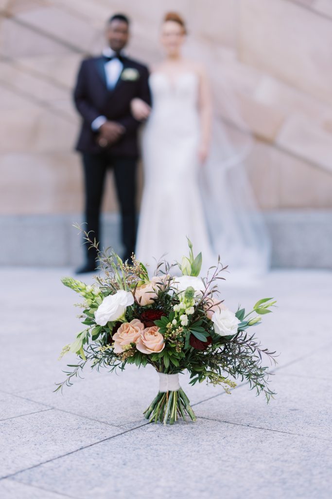 A bridal bouquet stands on the ground in Anzac Square Brisbane, featuring a sophisticated palette of rich burgundy and contrasting soft blush roses, chincherinchee and white lisianthus. The textured mix of greenery includes andromeda buddy the feath