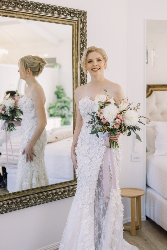 A smiling woman in a white wedding dress holding a bouquet of flowers, standing in front of a large mirror in a bright room.