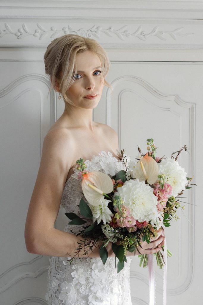 A bride in a lace wedding dress holding a bouquet of white and pink flowers, standing in front of an ornate white wall.