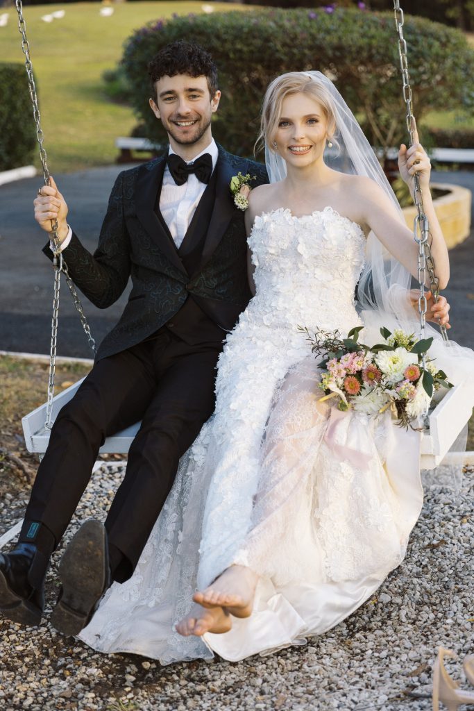 A newlywed couple sitting on a swing in a park, with the bride in a white wedding gown holding a bouquet and the groom in a tuxedo, both smiling.