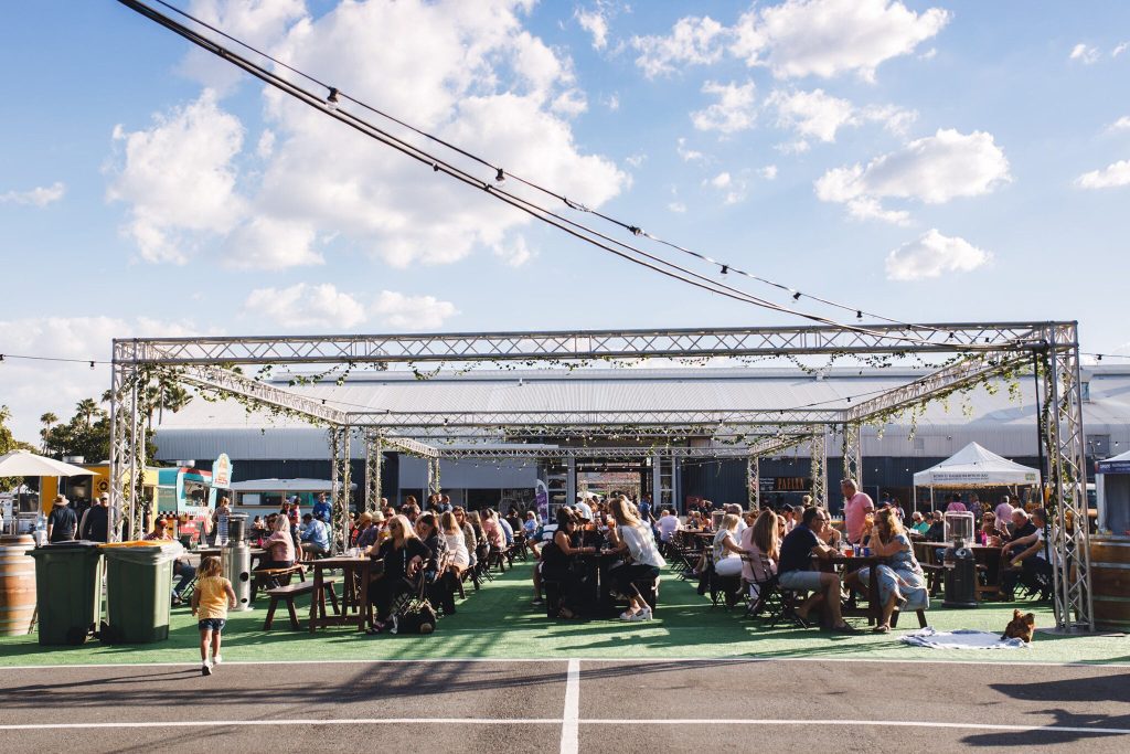 Outdoor food festival with many people sitting at tables under a metal frame canopy, with a few tents and food trucks in the background, on a bright day with a blue sky and scattered clouds.