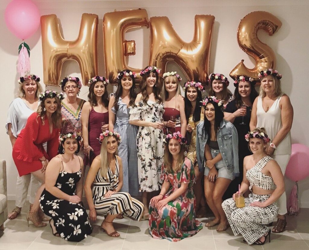 Group of women celebrating a bachelorette party, wearing flower crowns, in front of large gold balloons spelling "HEY S" with pink balloons on the sides.
