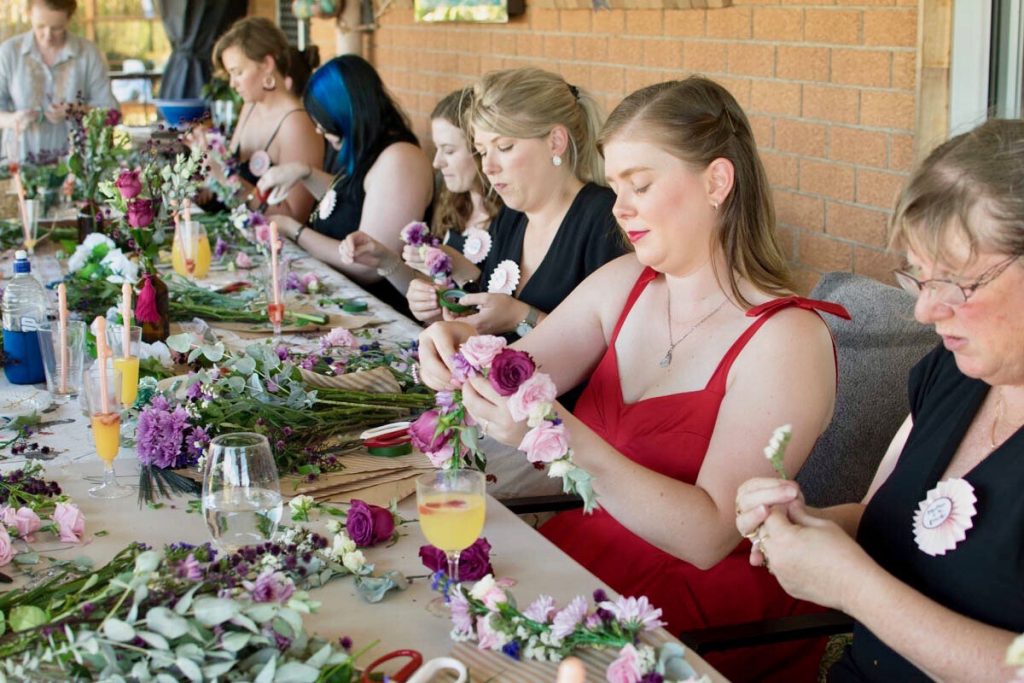 Women gather around a table creating floral arrangements with pink, purple, and white flowers
