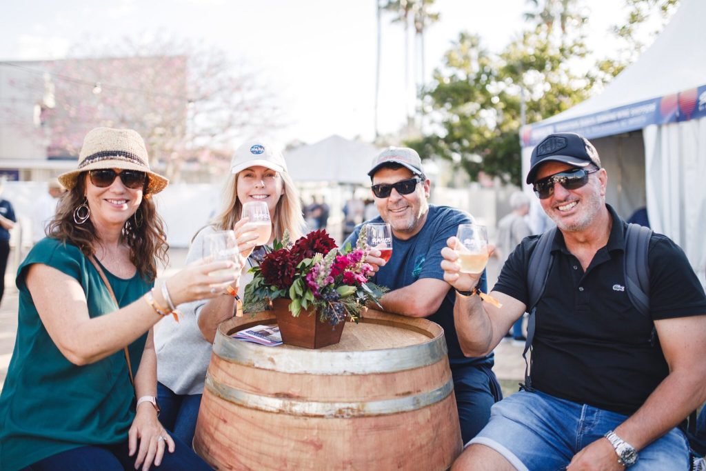 Four adults are enjoying drinks together at an outdoor event, sitting around a wooden barrel with a floral centerpiece, smiling at the camera.