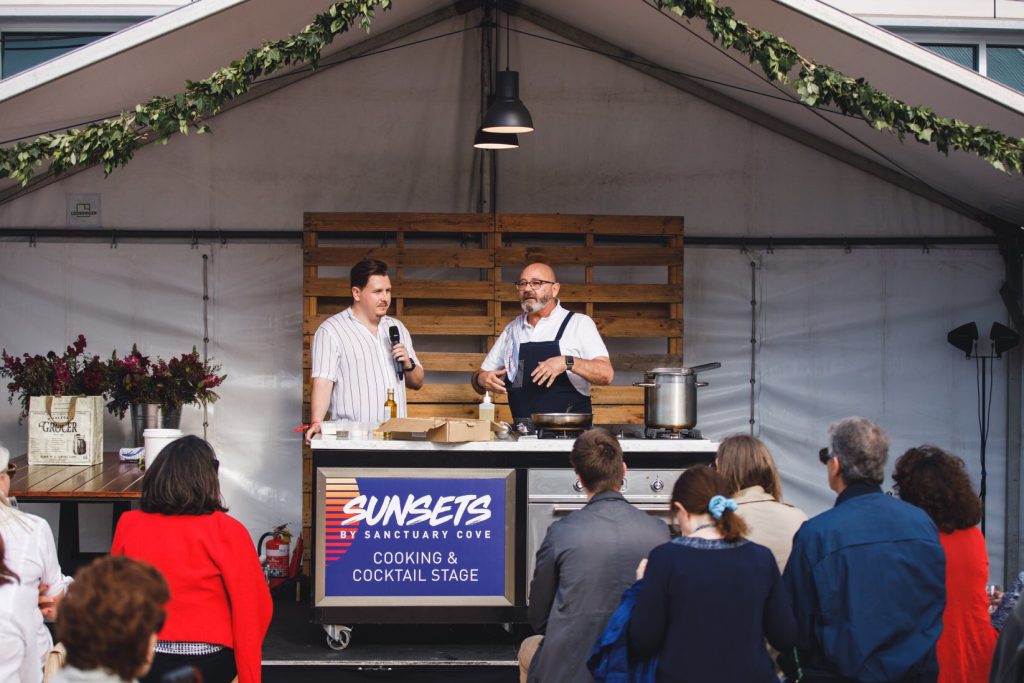 A cooking demonstration on a stage with two men, one with a microphone and the other wearing an apron, talking to an audience at an outdoor event called 'Sunsets by Sanctuary Cove'.