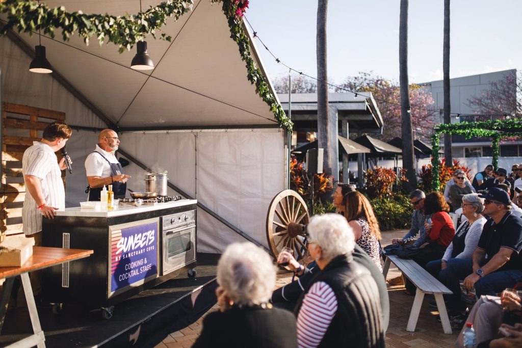 A cooking demonstration on an outdoor stage with an audience seated on benches. Two men are cooking, one speaking into a microphone, at Sunset by Sanctuary Cove with a sign indicating a cooking and cocktail stage. The scene is sunny with trees and fe