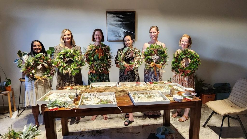 Six women standing behind a table creating floral wreaths, with craft supplies and greenery on the table, in a home or studio setting.