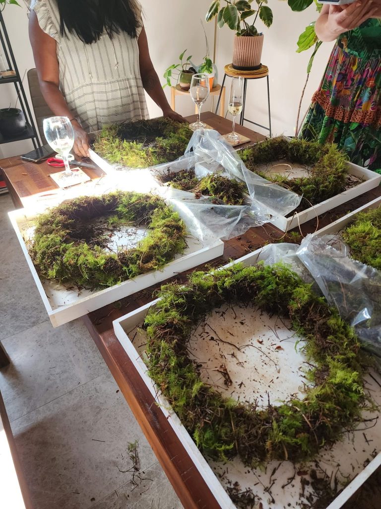 People assembling moss wreaths on white trays on a wooden table, with plants and glasses of wine nearby.