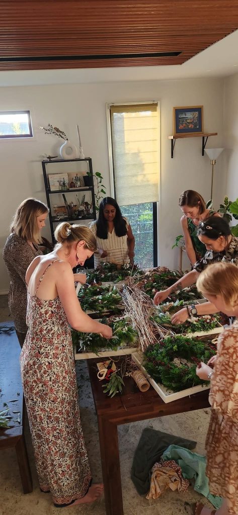 Group of women making holiday wreaths around a table with greenery, ribbons, and craft supplies in a bright room.