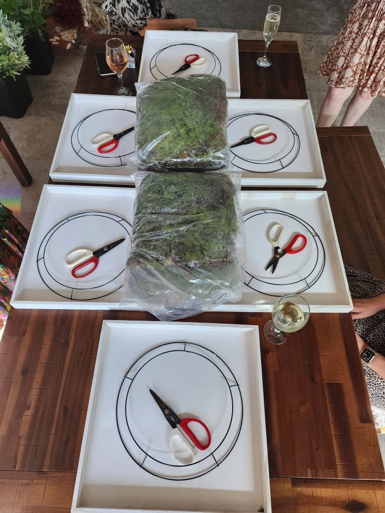 Table set with white trays, each with a pair of scissors and a lamb slice, and two large plastic bags filled with moss or greenery, on a wooden table with drinks and people around.