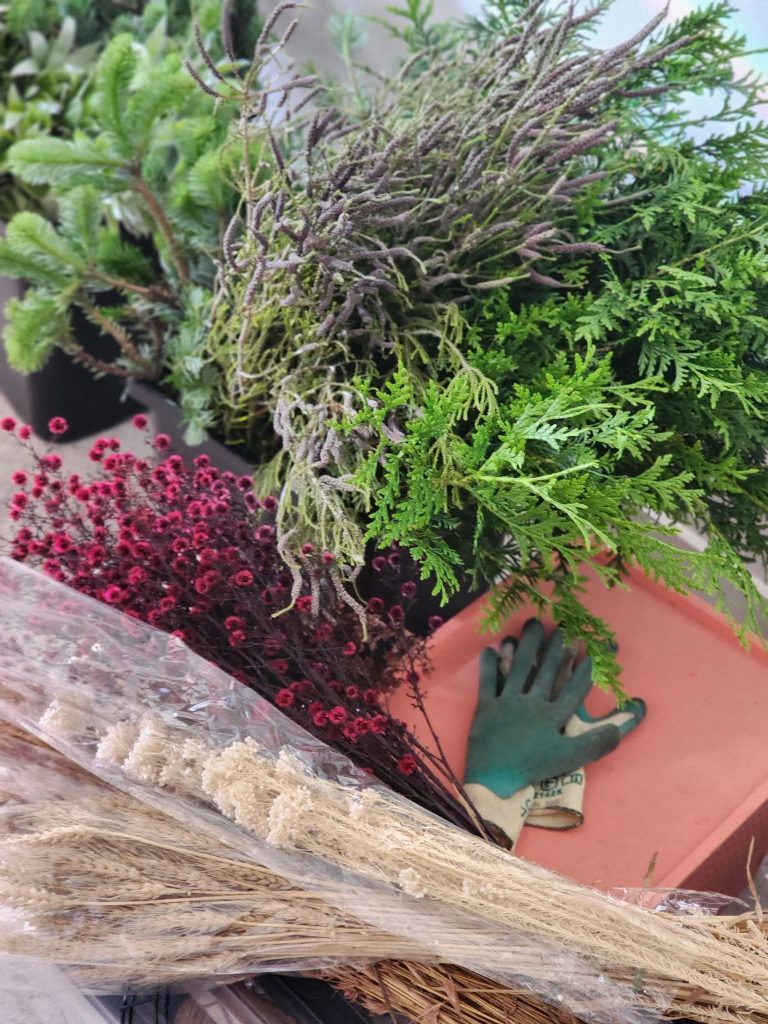A variety of potted plants, including ferns, lavender, and other greenery, with a gardening glove in a pink container and a bouquet of dried flowers and pink waxflower in the foreground.