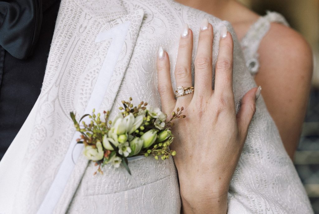 Close-up of a wedding ring on a woman's finger resting on her wedding dress, with a corsage of white flowers attached to her dress.
