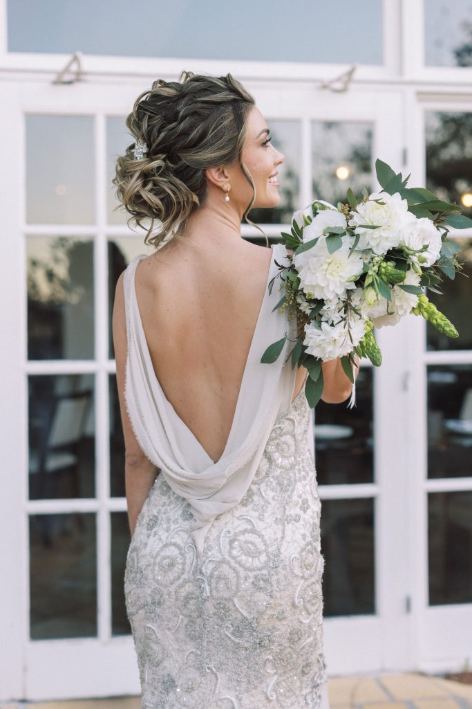 Bride wearing a white wedding dress with a low back, holding a bouquet of white flowers and greenery, standing in front of glass doors.