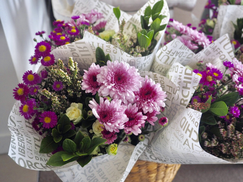 Bouquet of pink chrysanthemums, yellow roses, purple daisies, and small white flowers wrapped in decorative paper with words, placed in a wicker basket.