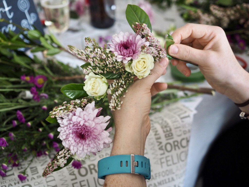 Person arranging a floral boutonniere with pink and white flowers on a table with other flowers and supplies.