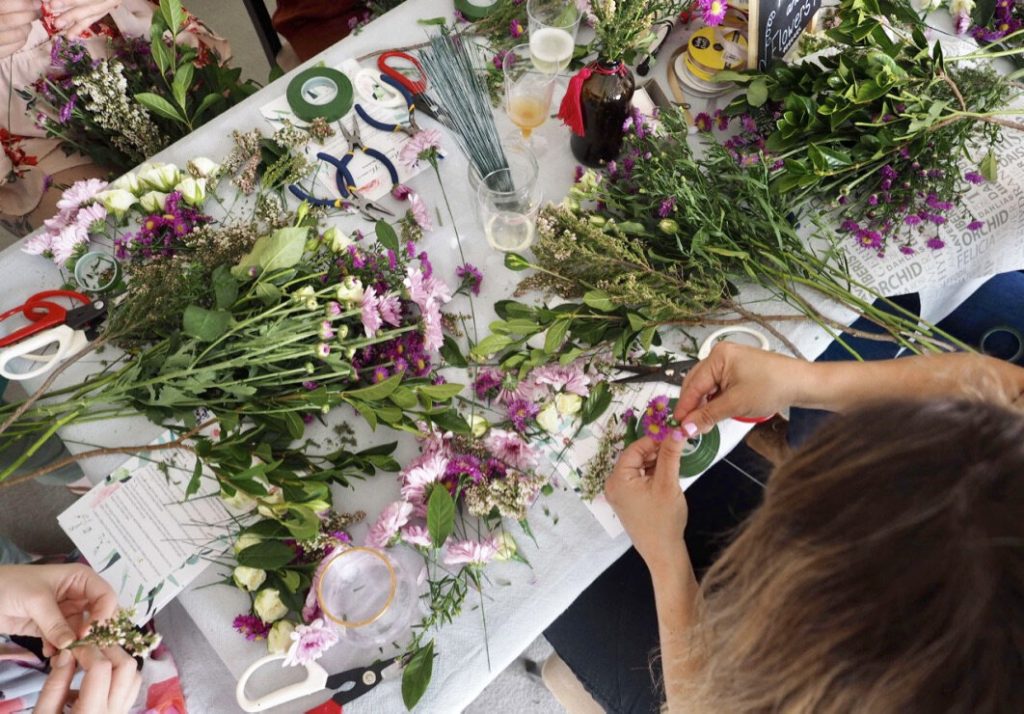 People arranging and creating floral bouquets with various flowers, greenery, scissors, tape, and other floral arranging tools on a white table.
