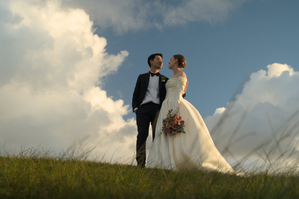 Wedding portrait featuring a couple stood on the Lennox Head headlands, showcasing a beautiful bridal bouquet created by a Brisbane florist Hart and Flowers amidst rolling hills and a cloud-filled sky.