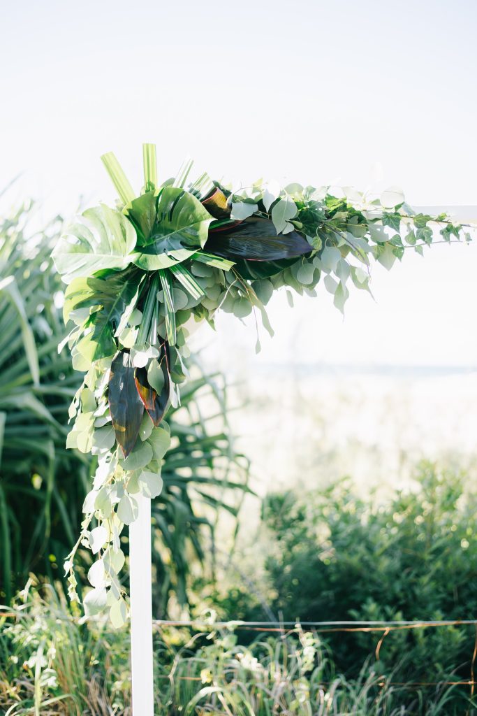 A lush and tropical wedding arch adorned with a vibrant mix of greenery. The arch features large, dramatic leaves, including monstera leaves, eucalyptus, and other tropical foliage, creating a bold and eye-catching statement piece. The arch is set ag