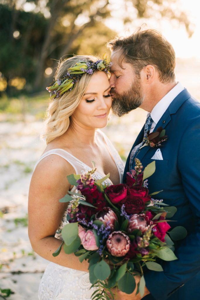 A vibrant and dramatic bouquet featuring a mix of deep burgundy roses, vibrant proteas, and lush greenery. The arrangement showcases a bold color palette and a mix of textures, creating a captivating and eye-catching display. The overall effect is on