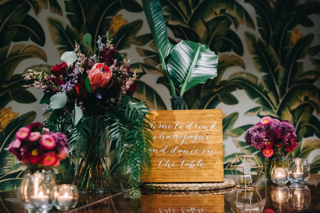 A vibrant wedding reception setting with a tropical theme atop a piano. The piano is adorned with a variety of colorful flowers, including proteas and chrysanthemums, arranged in vases and jars. A wooden sign with the phrase "Time to drink champagne