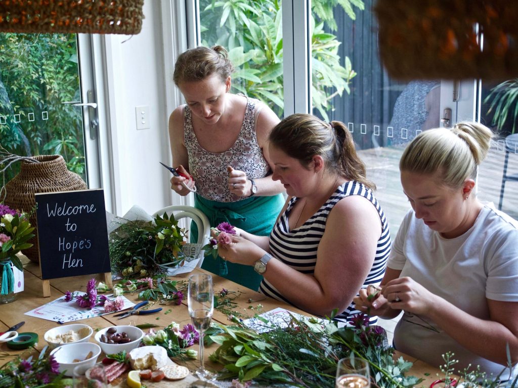 Three women making flower arrangements at a table with various flowers, greenery, and tools, next to a window with outdoor greenery view, and a sign that reads 'Welcome to Hope's Hen'.