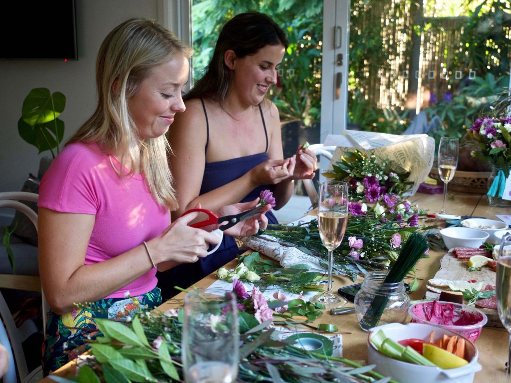 Two women, one with blonde hair in a pink shirt and another with brown hair in a blue dress, are sitting at a table assembling floral arrangements. The table is filled with flowers, tools, and drinks, and they are smiling while working.