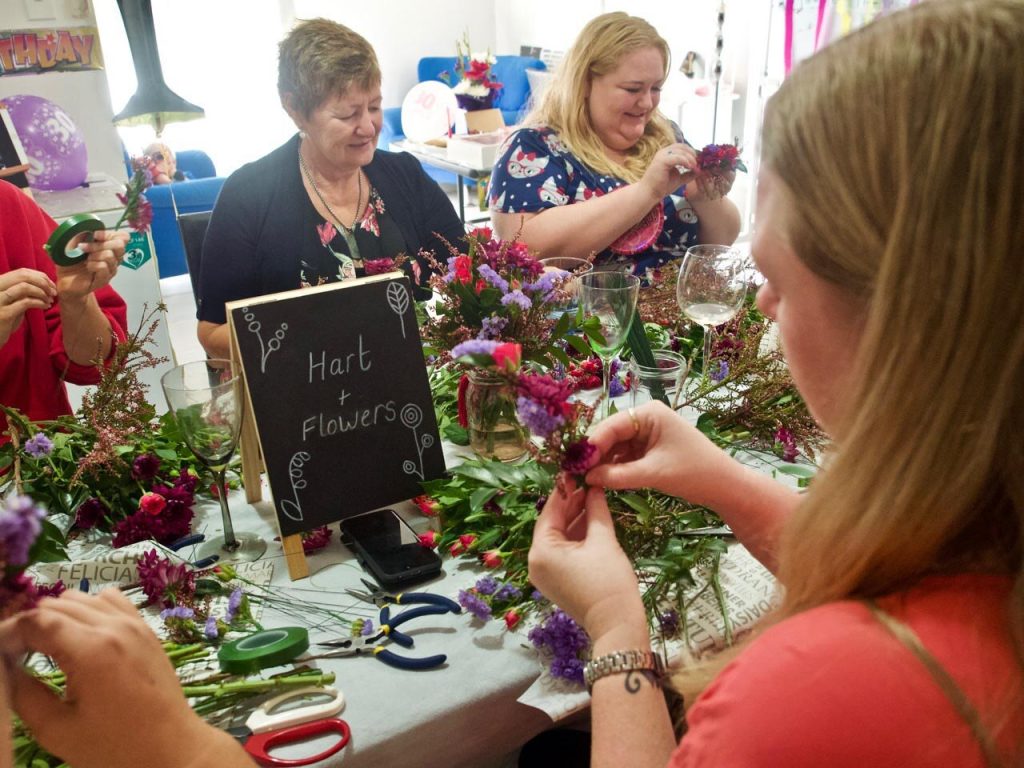 Four women gathered around a table arranging colorful flowers for a floral craft activity, with a blackboard sign that reads 'Hart + Flowers' and various floral tools on the table.