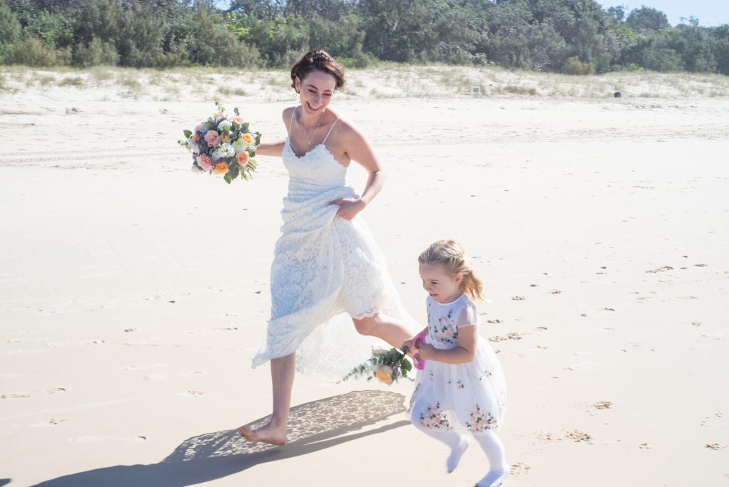 A bride playfully runs on a Gold Coast beach alongside her young flower girl holding their bouquets featuring peach roses, fragrant white stock, limonium and textured eucalyptus greenery created by Hart and Flowers.