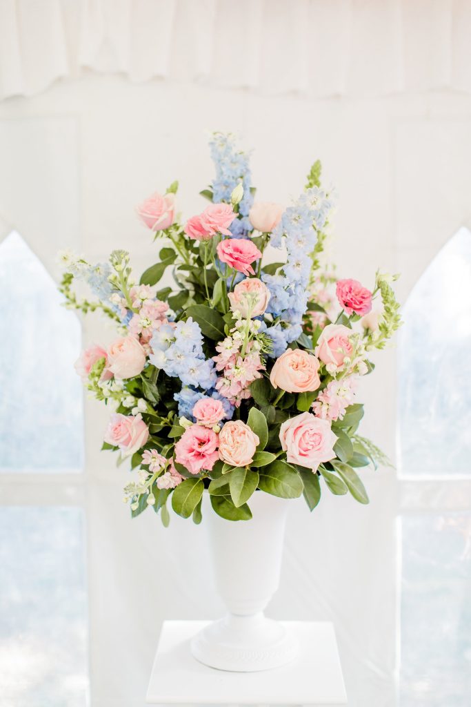 A colorful bouquet of pink, peach, and blue flowers in a white vase on a white pedestal.