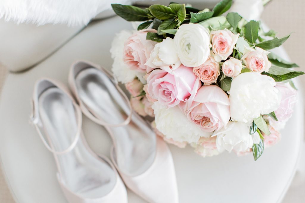 A pair of pale pink high-heeled shoes and a bouquet of pink and white flowers on a white surface.